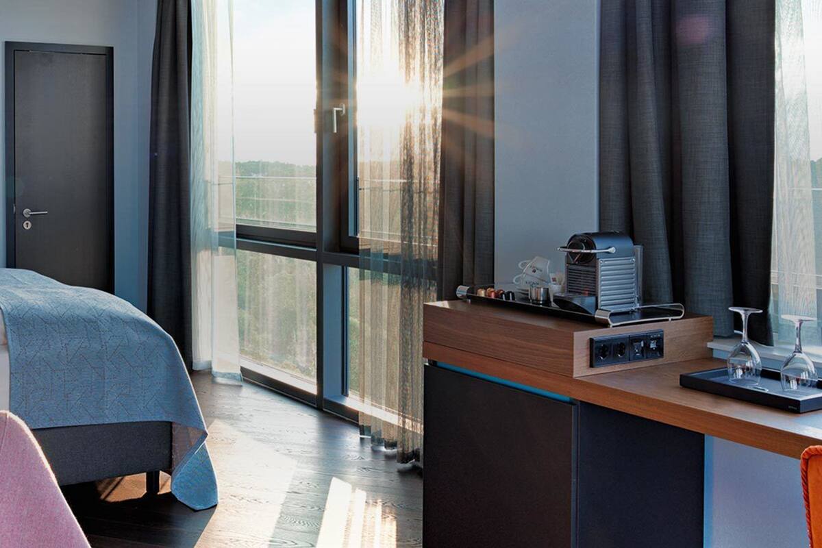 A modern hotel room with a bed, bedside lamps, a desk with a coffee maker, and a large window with a view.