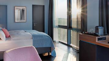 A modern hotel room with a bed, bedside lamps, a desk with a coffee maker, and a large window with a view.