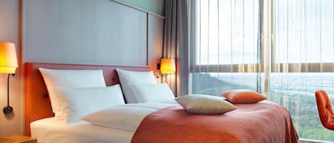 A hotel room with a large bed, two bedside lamps, a view of the outdoors through the window, and a wooden nightstand with a lamp.