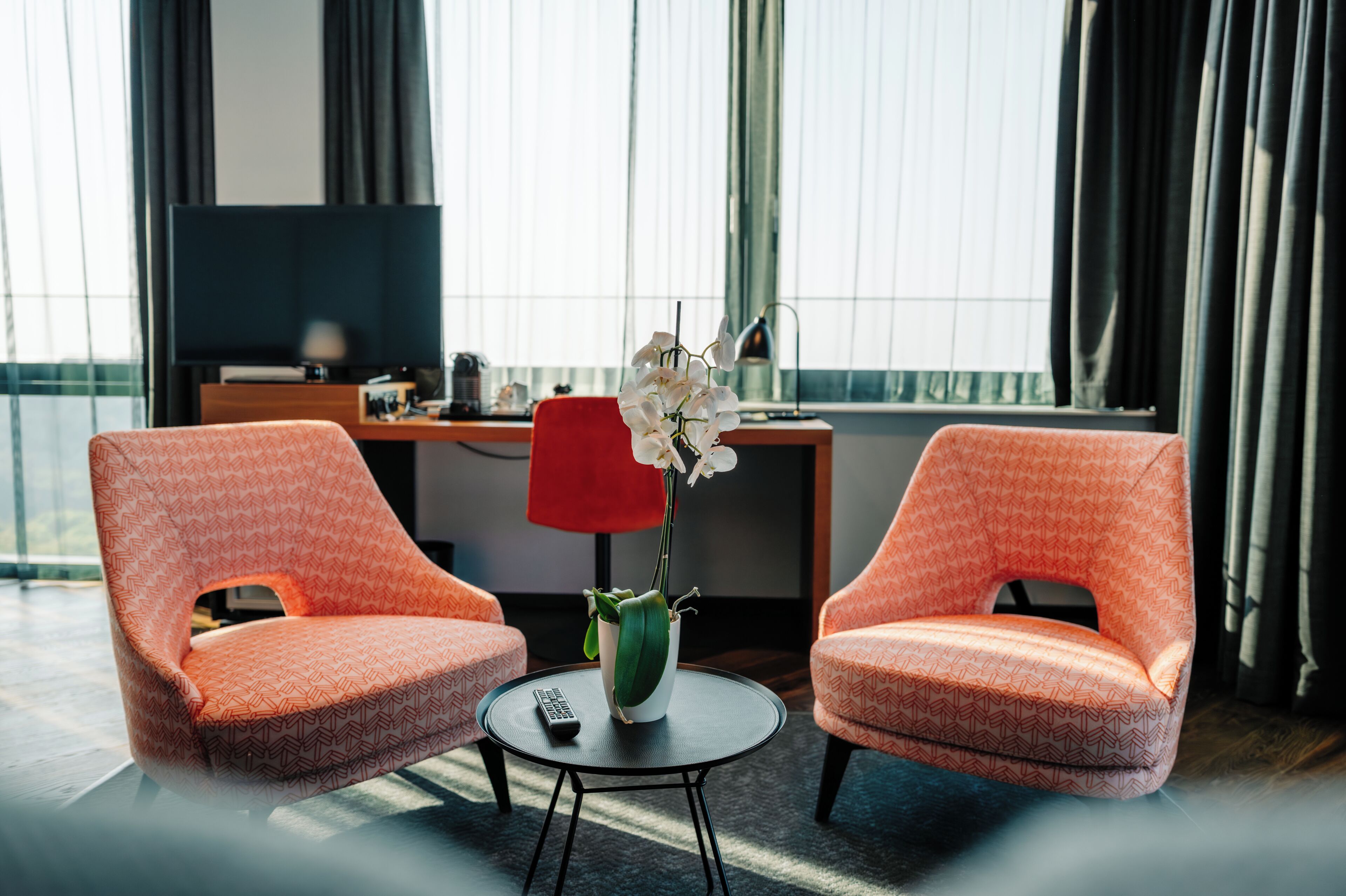 Une chambre d’hôtel avec deux fauteuils à motifs, une petite table ronde avec une télécommande, un vase de fleurs blanches et un bureau avec une lampe et un téléviseur.