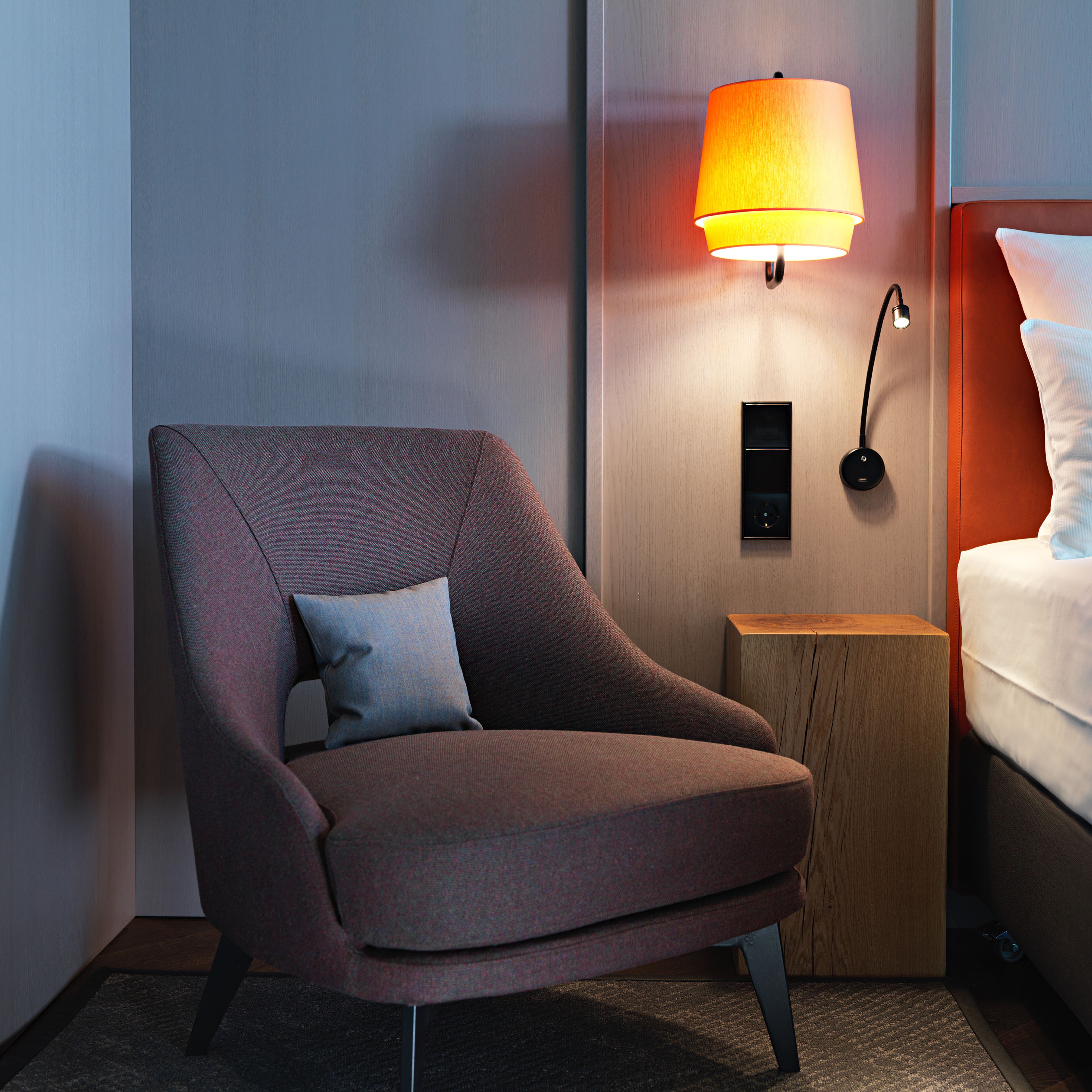 Une chambre d’hôtel moderne, dotée d’un fauteuil, d’une table de chevet avec une lampe et d’un lit recouvert de linge de maison blanc.