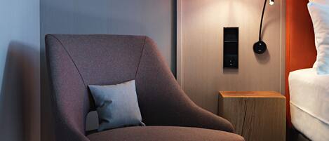 Une chambre d’hôtel moderne, dotée d’un fauteuil, d’une table de chevet avec une lampe et d’un lit recouvert de linge de maison blanc.