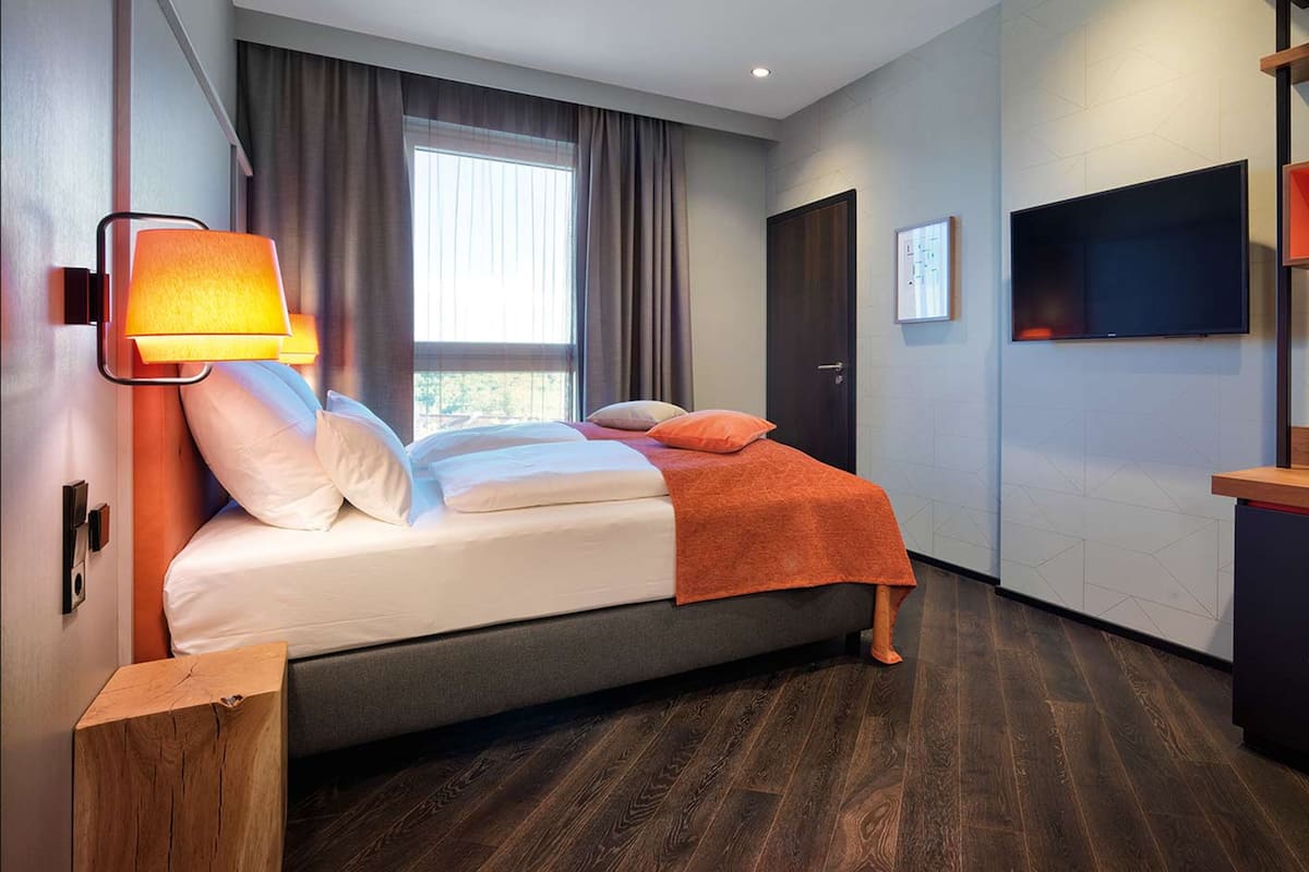 A modern hotel room with a bed, a nightstand, a TV, and a window with curtains.
