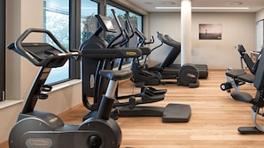 Een sportschool met meerdere loopbanden en elliptische trainers, grote ramen en een houten vloer.