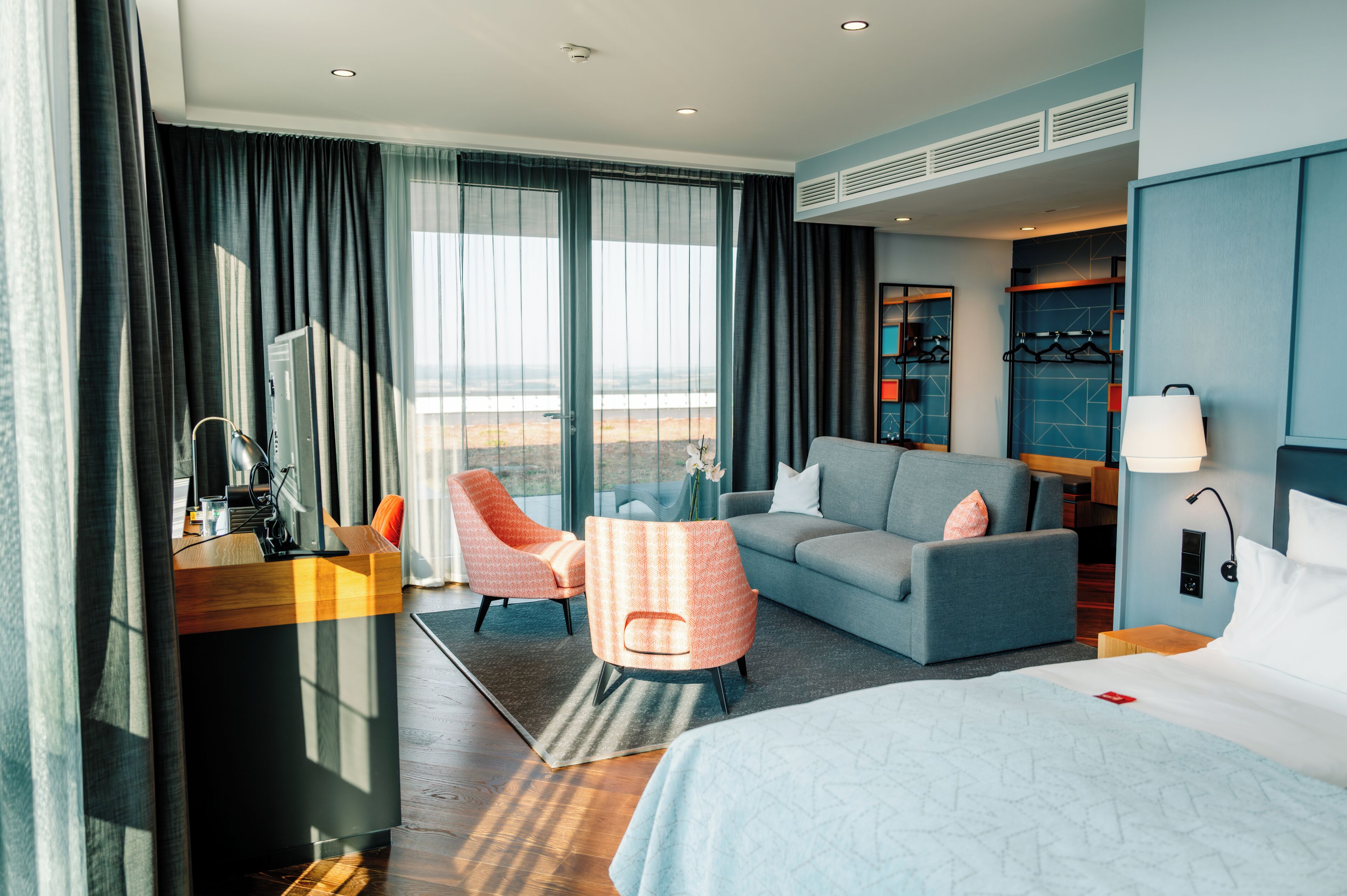 A hotel room with a bed, a sofa, a desk, and a chair. There is a view of the outdoors through the windows.