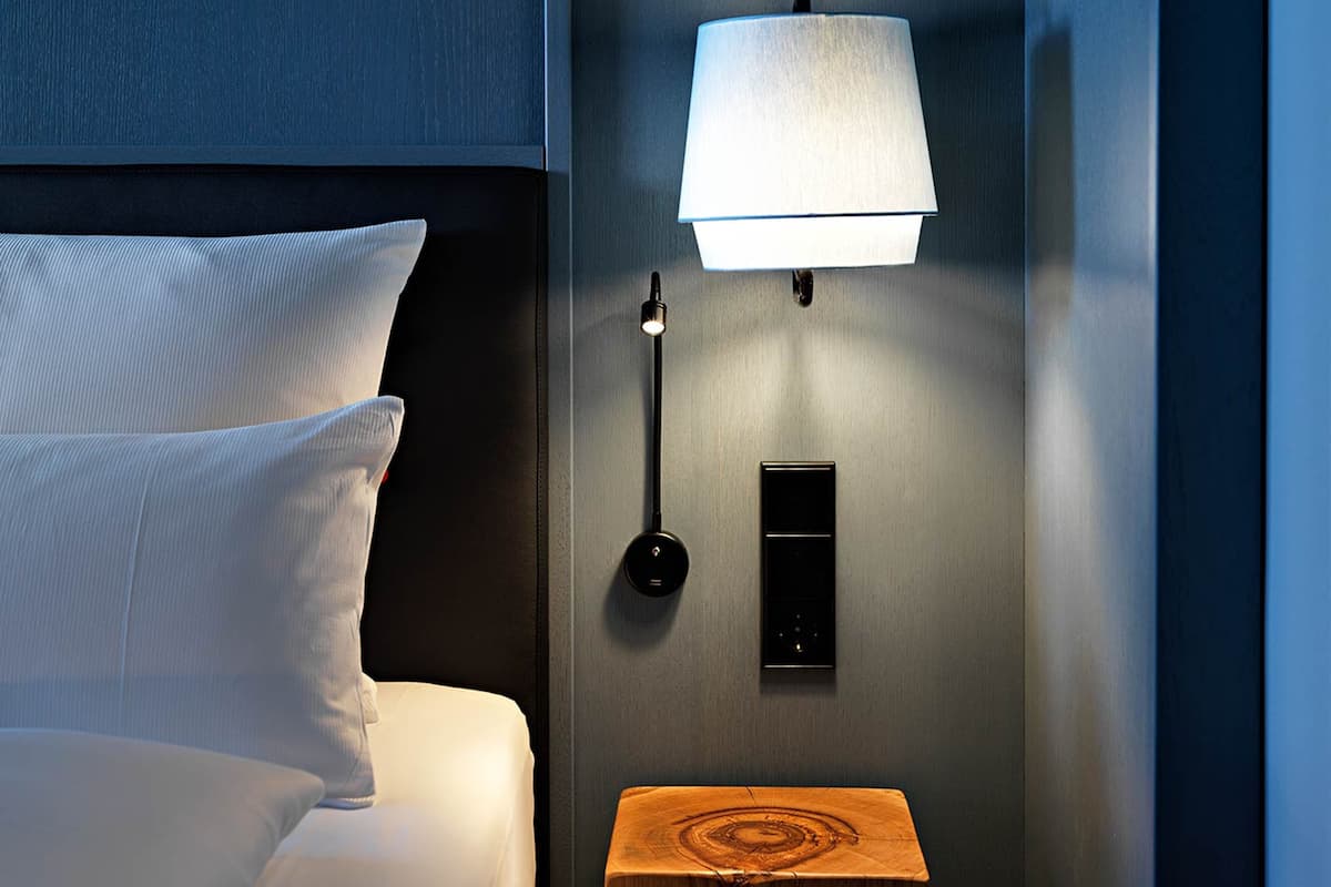 A modern hotel room with a bed, a bedside table, a lamp, and a wall-mounted control panel.