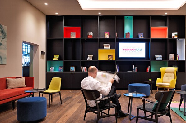 A modern hotel lobby with a large screen displaying the hotel name, a variety of seating options including a red sofa, yellow and black chairs, and a blue ottoman. - Panoramahotel Waldenburg (Waldenburg)