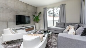 A modern living room with a flat-screen TV, grey sofas, a coffee table, and a large window with curtains.