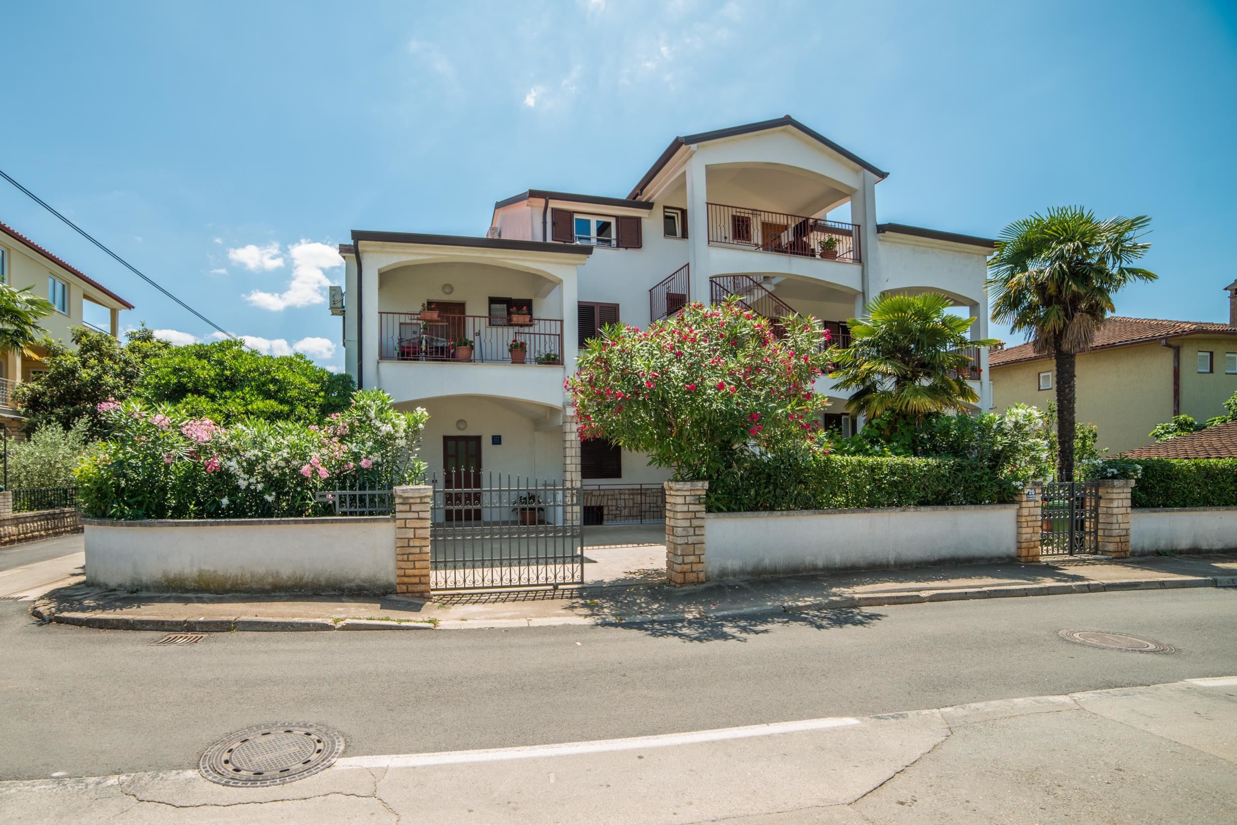 A two-story residential building with balconies, a gated entrance, and surrounding greenery.