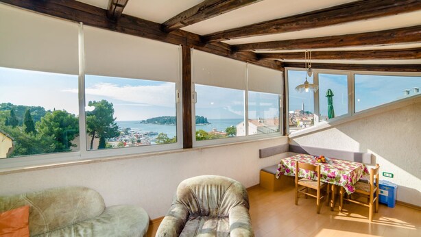 Una habitación con techo de madera, ventanas amplias con vistas a un pueblo y al mar, un sofá, una silla y una mesa con mantel.