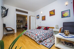 A modern bedroom with a bed, a desk, and a chair. There are paintings on the wall and a plant on the floor.