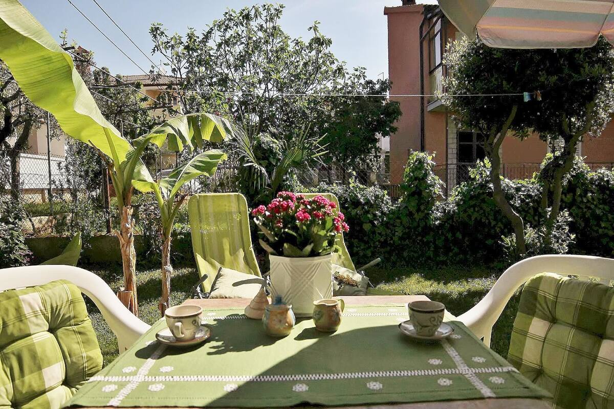 A patio with a table set for two, surrounded by lush greenery and a building in the background.