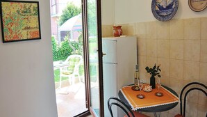 Quarto com mesa de jantar posta para dois, geladeira, planta em vaso e janela com vista para um jardim e prédios.