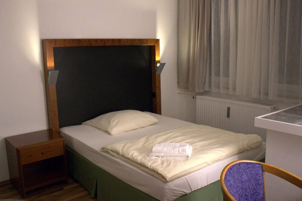 A single bed with a wooden headboard, a bedside table, a chair, and a window with curtains.