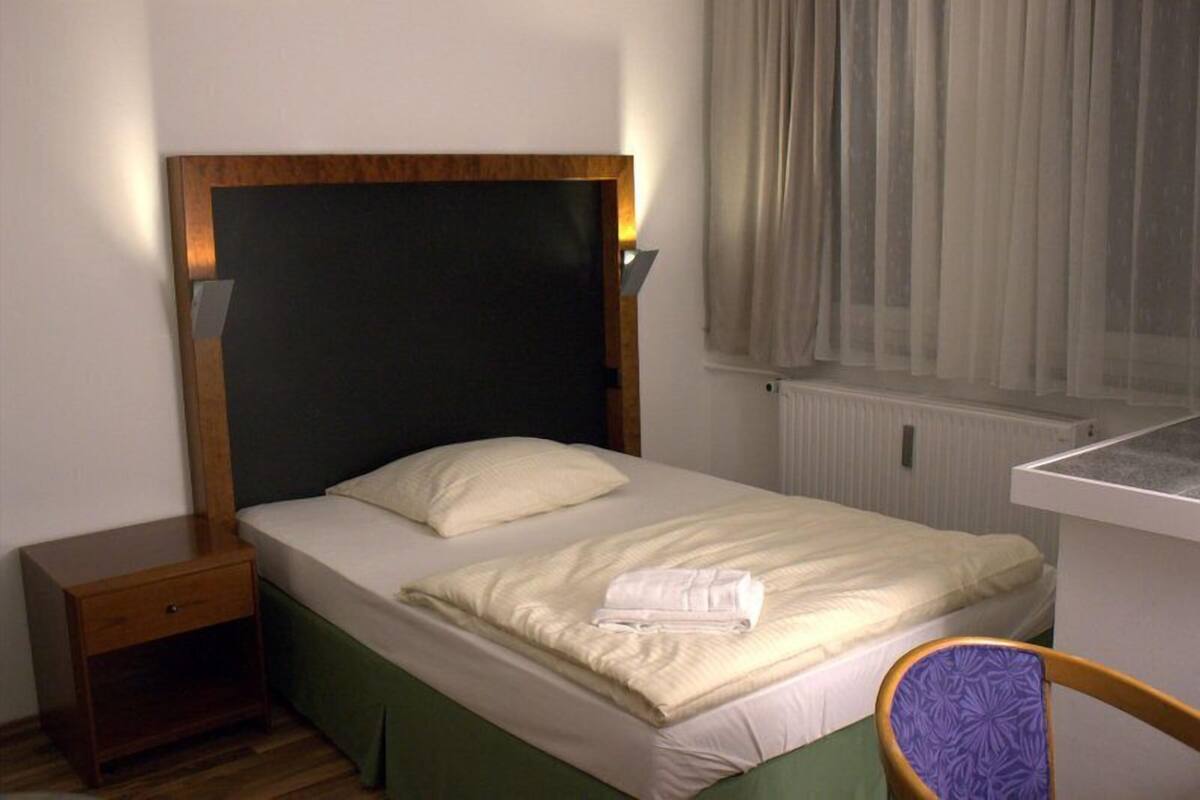 A single bed with a wooden headboard, a bedside table, a chair, and a window with curtains.