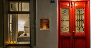 A hotel entrance with a red door, a visible room with a bed and a nightstand, and a small window with a view of the sky.