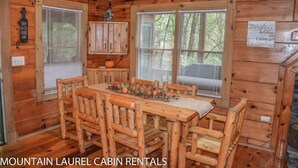 A wooden cabin interior with a dining table, chairs, a cabinet, and a window with blinds. - Chasing Rainbows (Mineral Bluff)