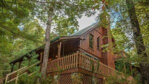A cabin with a wooden exterior, a balcony, and a stone foundation, surrounded by trees. - Chasing Rainbows (Mineral Bluff)