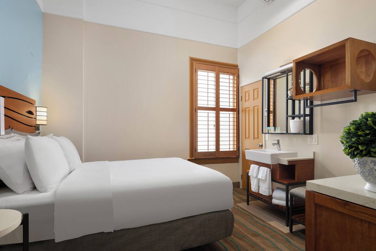 Habitación de hotel con cama, baño con lavamanos y espejo, y ventana con persianas.