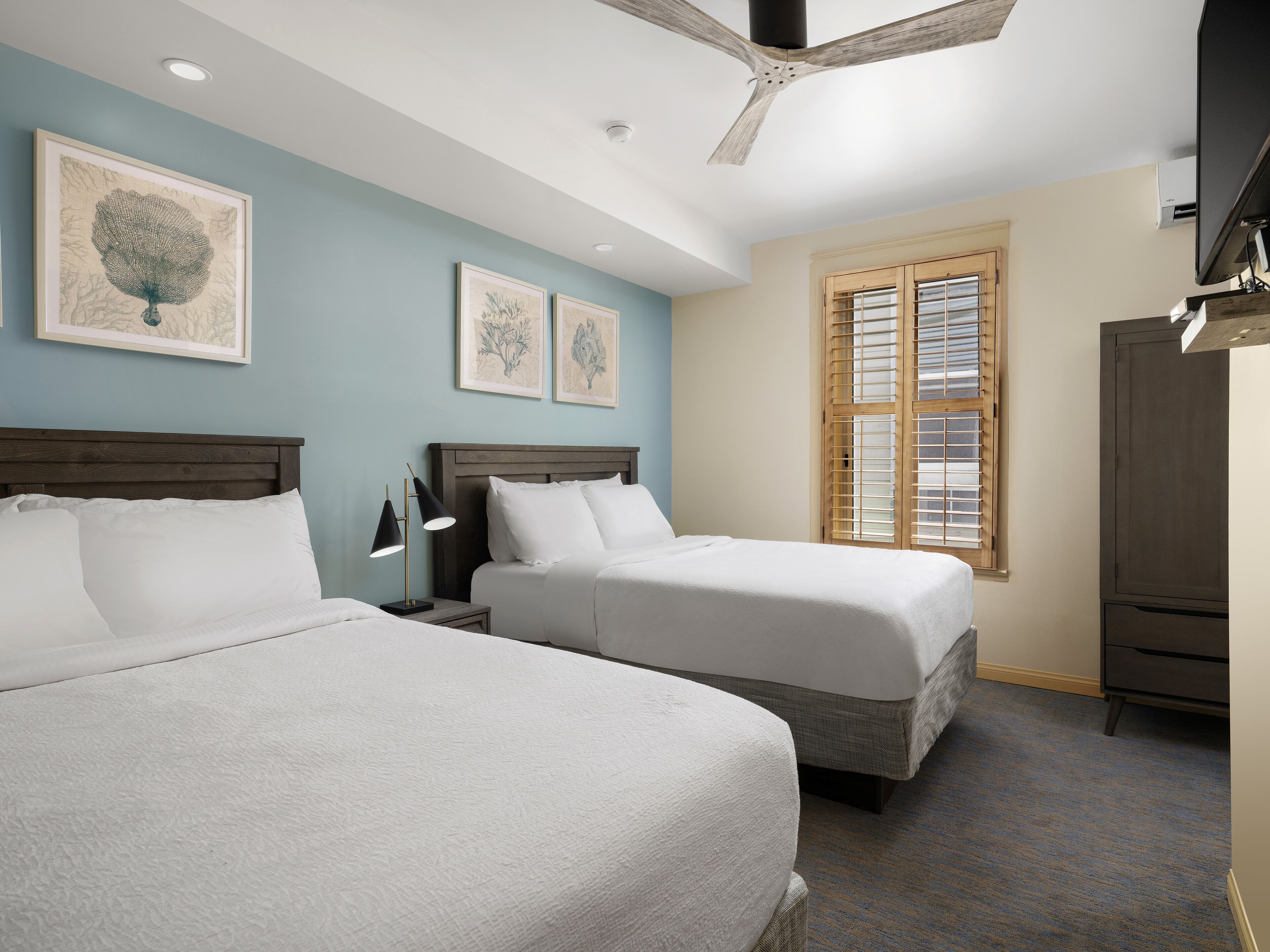 A hotel room with two beds, a ceiling fan, and artwork on the walls.