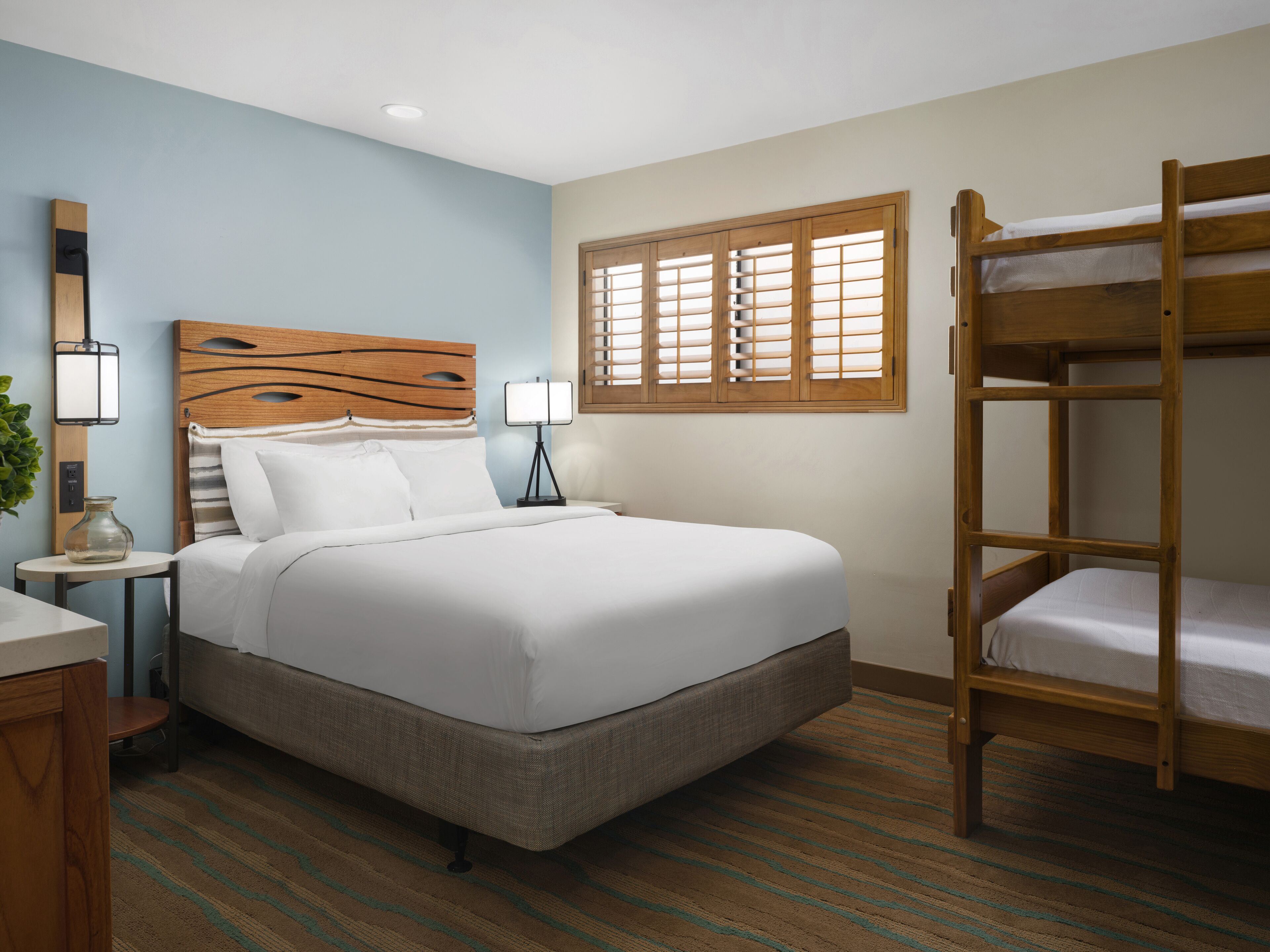 A hotel room with a large bed, a wooden bunk bed, a bedside table with lamps, and a window with blinds.