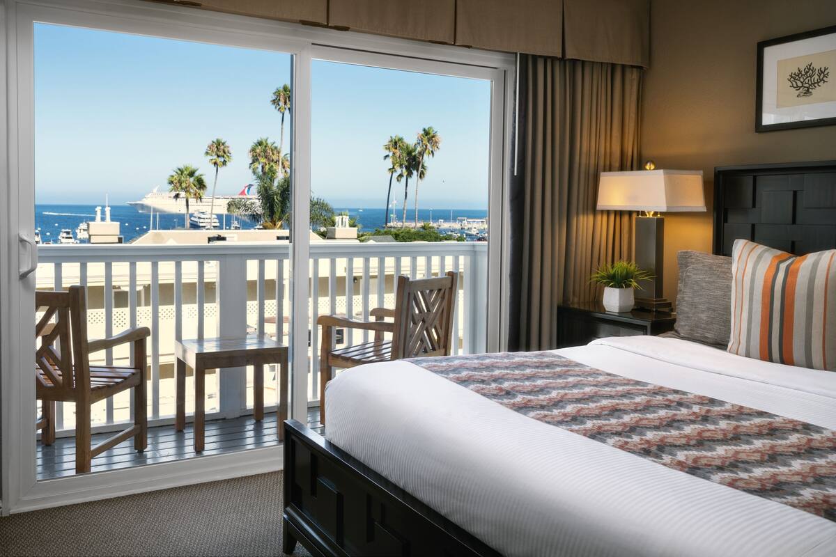 Un cuarto de hotel con balcón, una cama con cubrecama estampado, una mesita de noche con lámpara y vista al océano y palmeras.