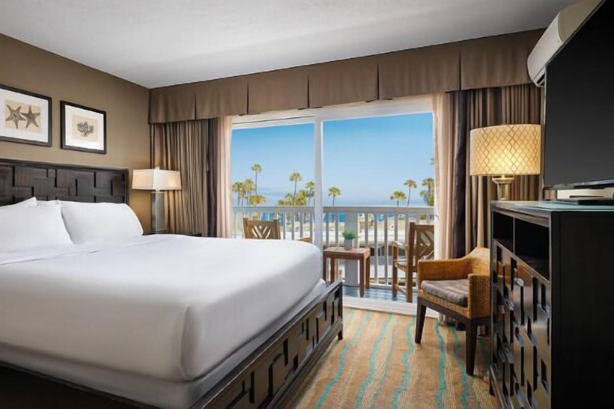 Habitación de hotel con una cama grande, un escritorio con televisor, y un balcón con vistas a palmeras y al océano.