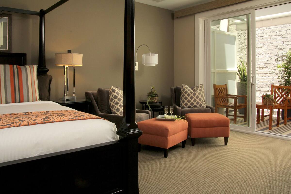 Habitación de hotel con cama con dosel, dos reposapiés, un sofá, una silla, una mesita con bandeja y una planta en maceta.