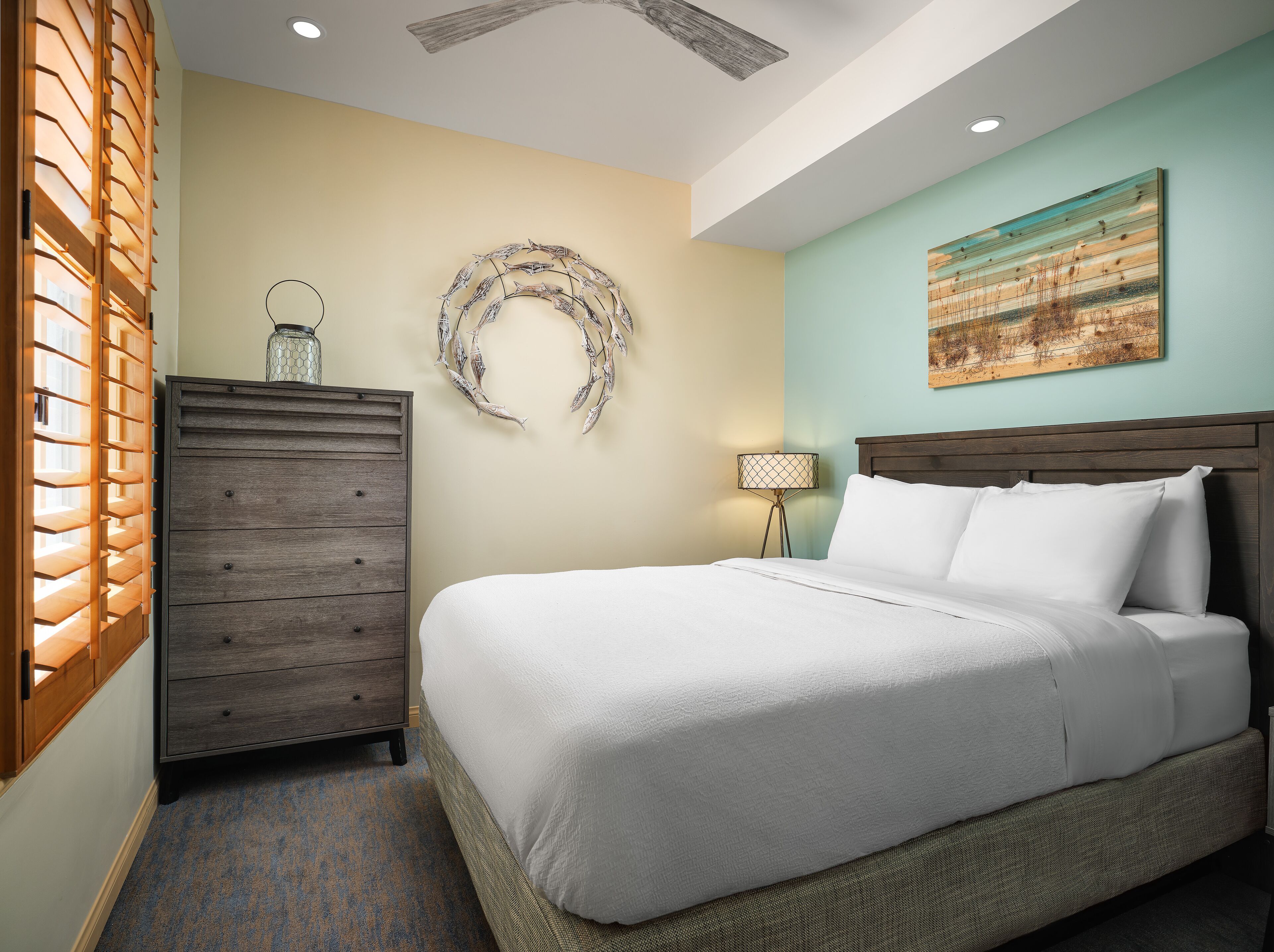 A neatly made bed with white linens, a wooden dresser, a wall-mounted circular fish decoration, and a painting above the bed.