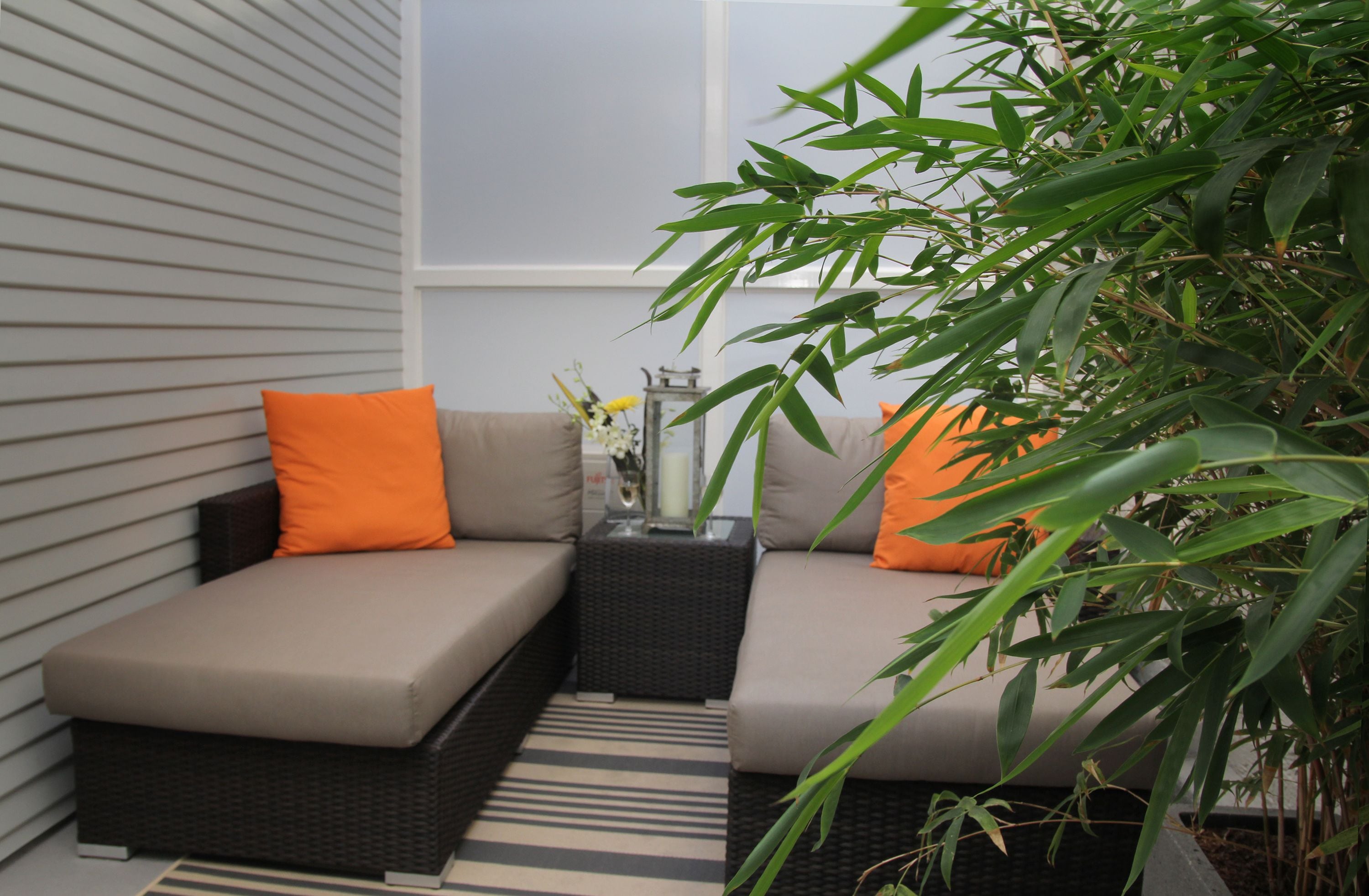 Een gezellig buitenverblijf met een comfortabele bank, oranje kussens, een kleine tafel met een vaas en een bamboeplant.