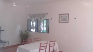 A dining area with a table covered by a white tablecloth, surrounded by four red chairs. There is a potted plant to the left of the table and a window with curtains above it. - Casa Candil, cozy and intimate Mayan corner (Pixoy)