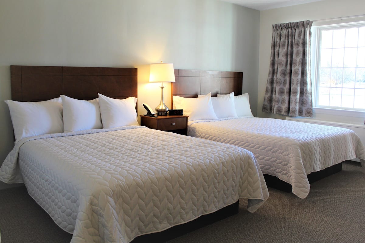Two queen beds with white bedding, a nightstand with a lamp, and a window with curtains.