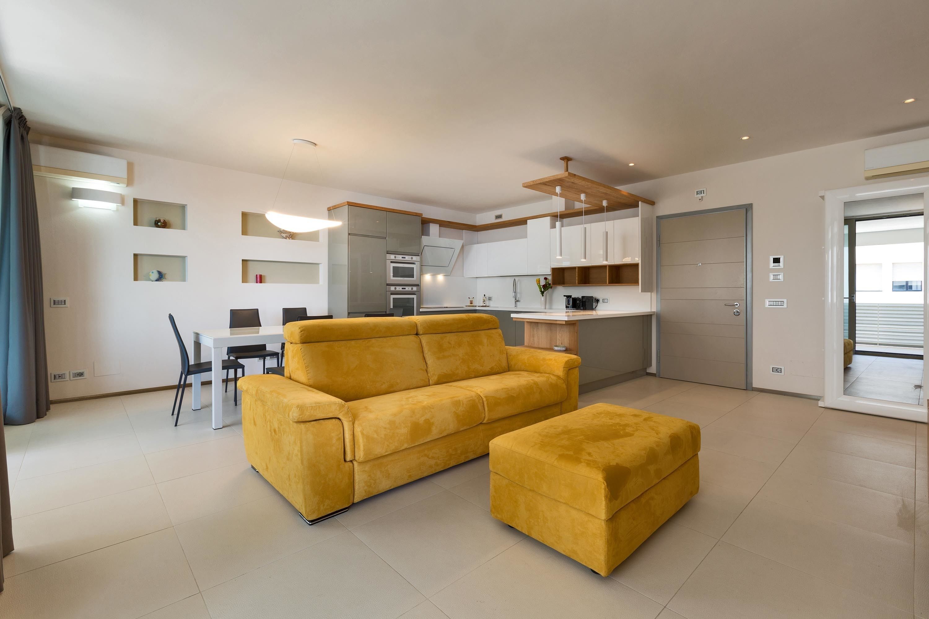 Een moderne woonkamer met een gele bank en poef, een eethoek met een witte tafel en stoelen, en een keuken met witte kasten en een houten plafond.