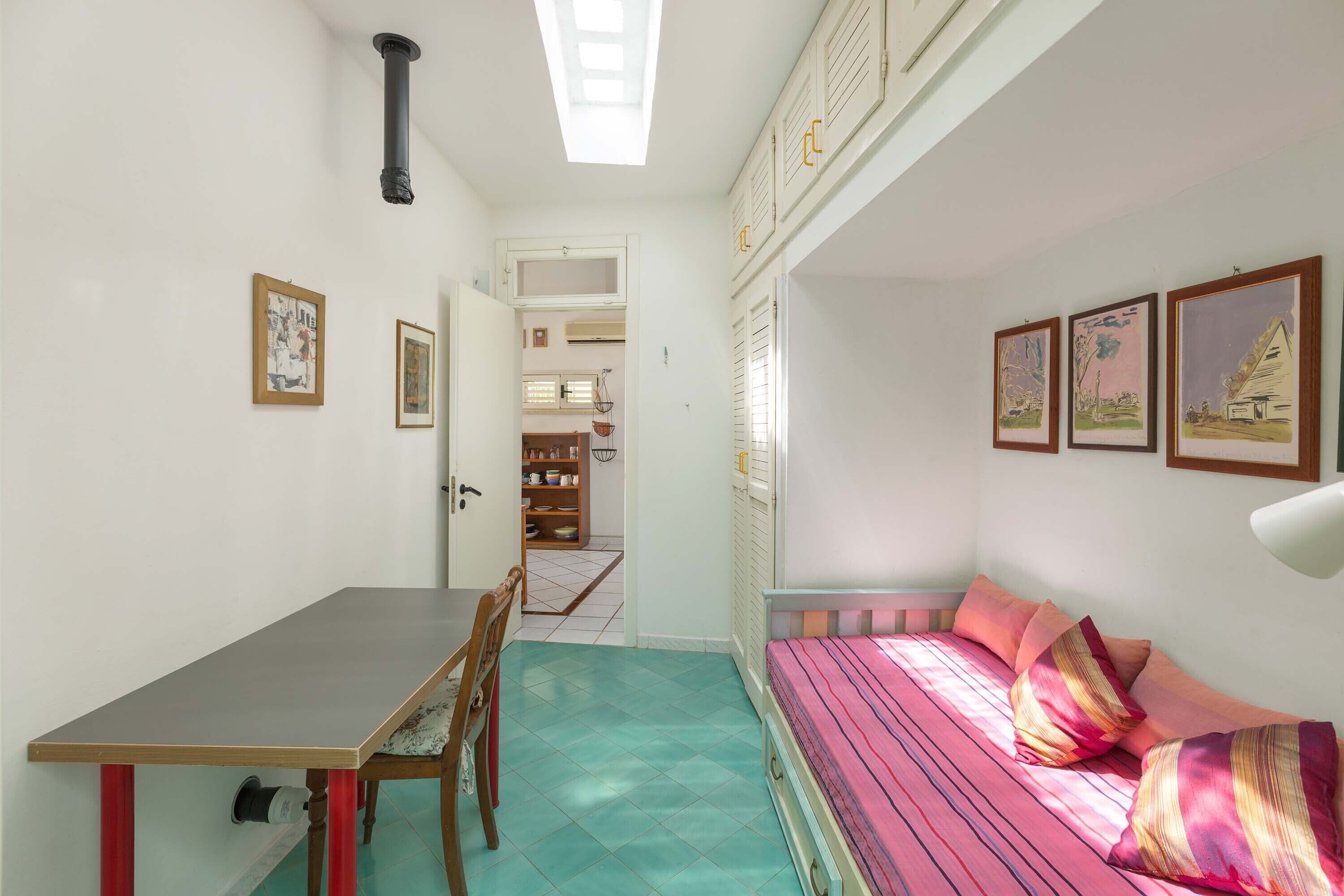 Une pièce avec un lit, une table et une chaise. Des tableaux encadrés ornent le mur.