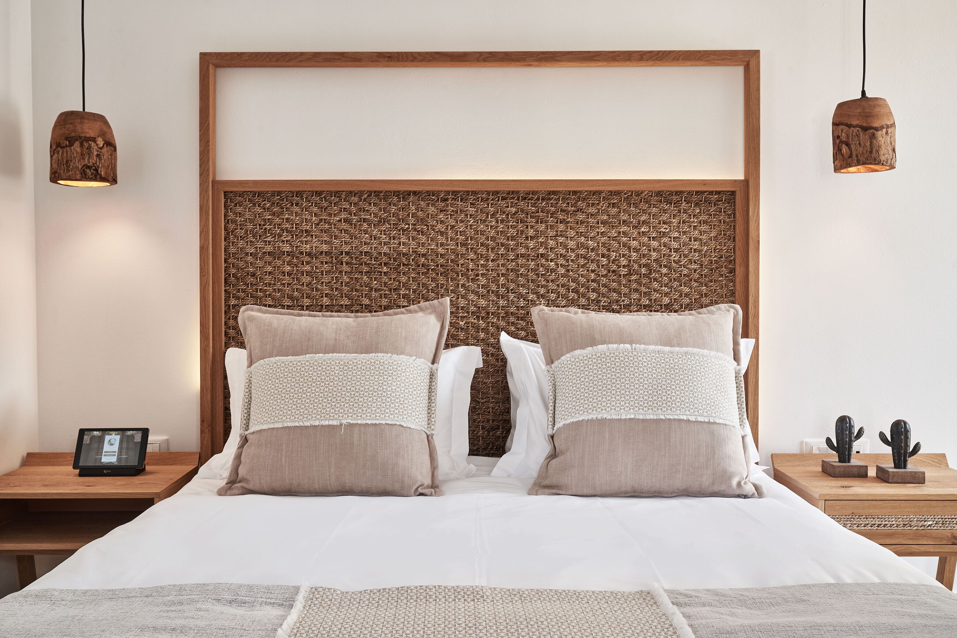 Una cama bien tendida con un cabecero tejido, dos mesitas de noche con una tableta y una decoración de cactus, y dos lámparas colgantes.