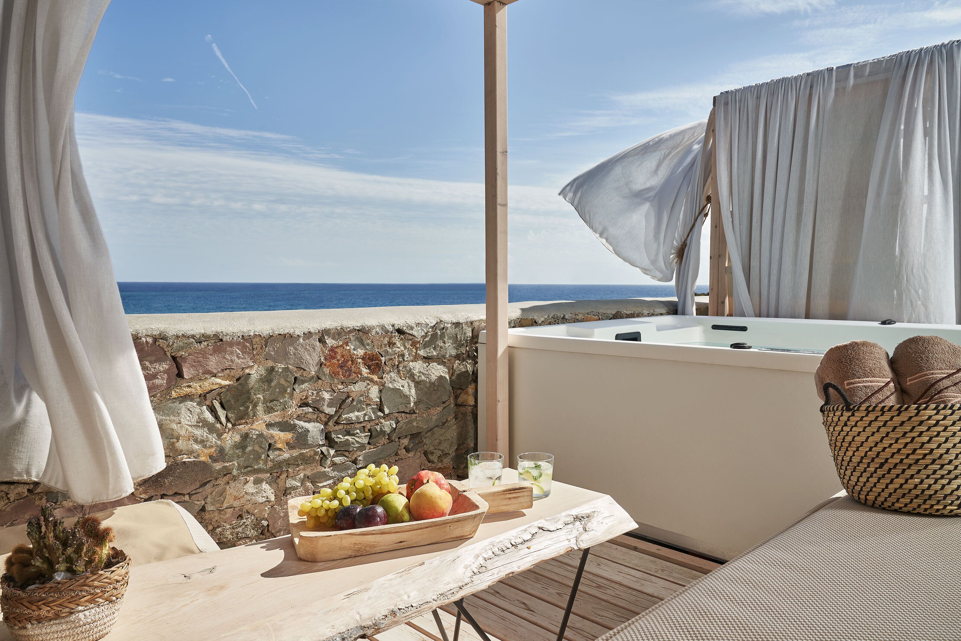 A balcony with a stone wall, a table with fruits and a basket, and a view of the sea.
