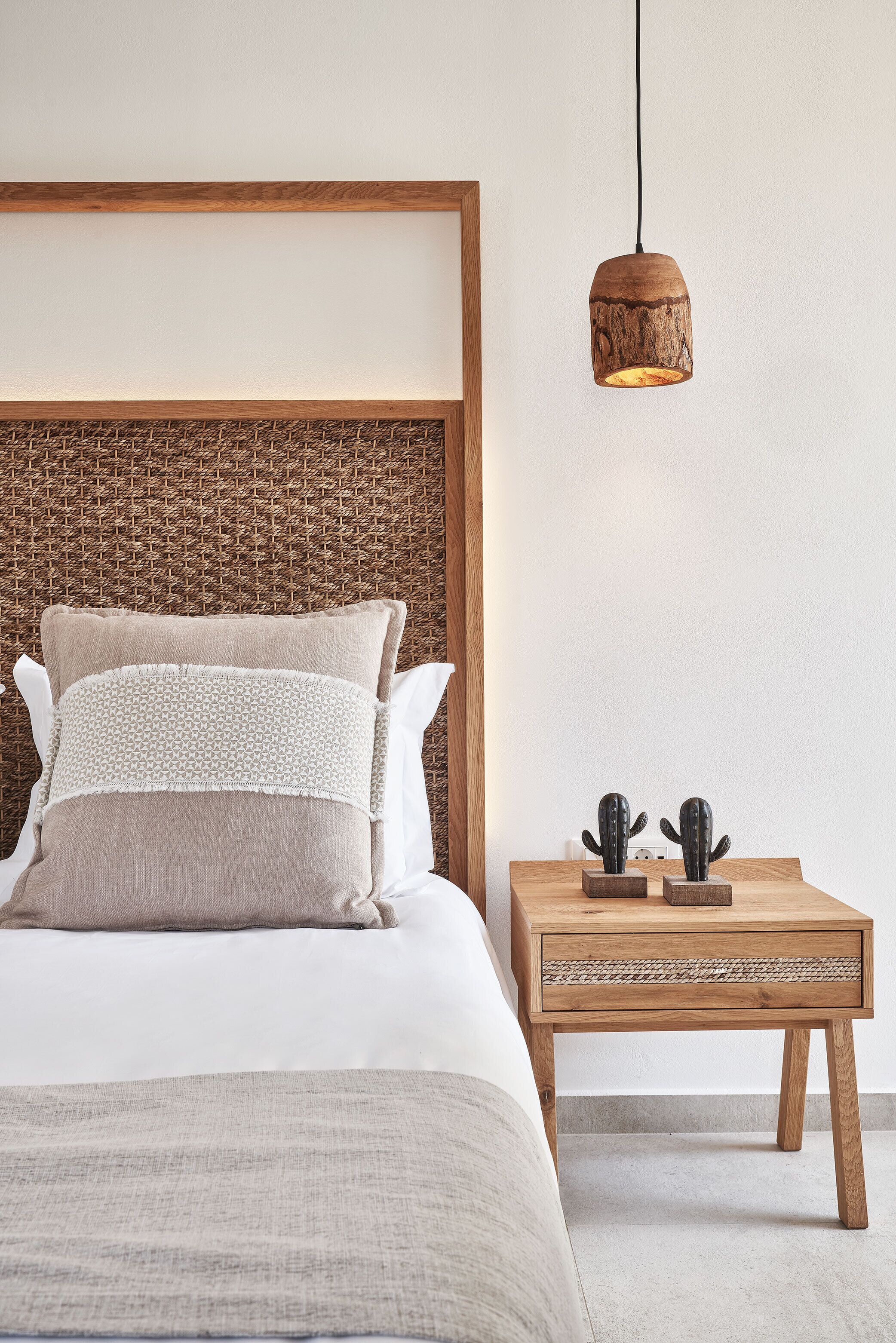 A neatly made bed with a wooden headboard, a bedside table with a woven texture, and a hanging pendant light.