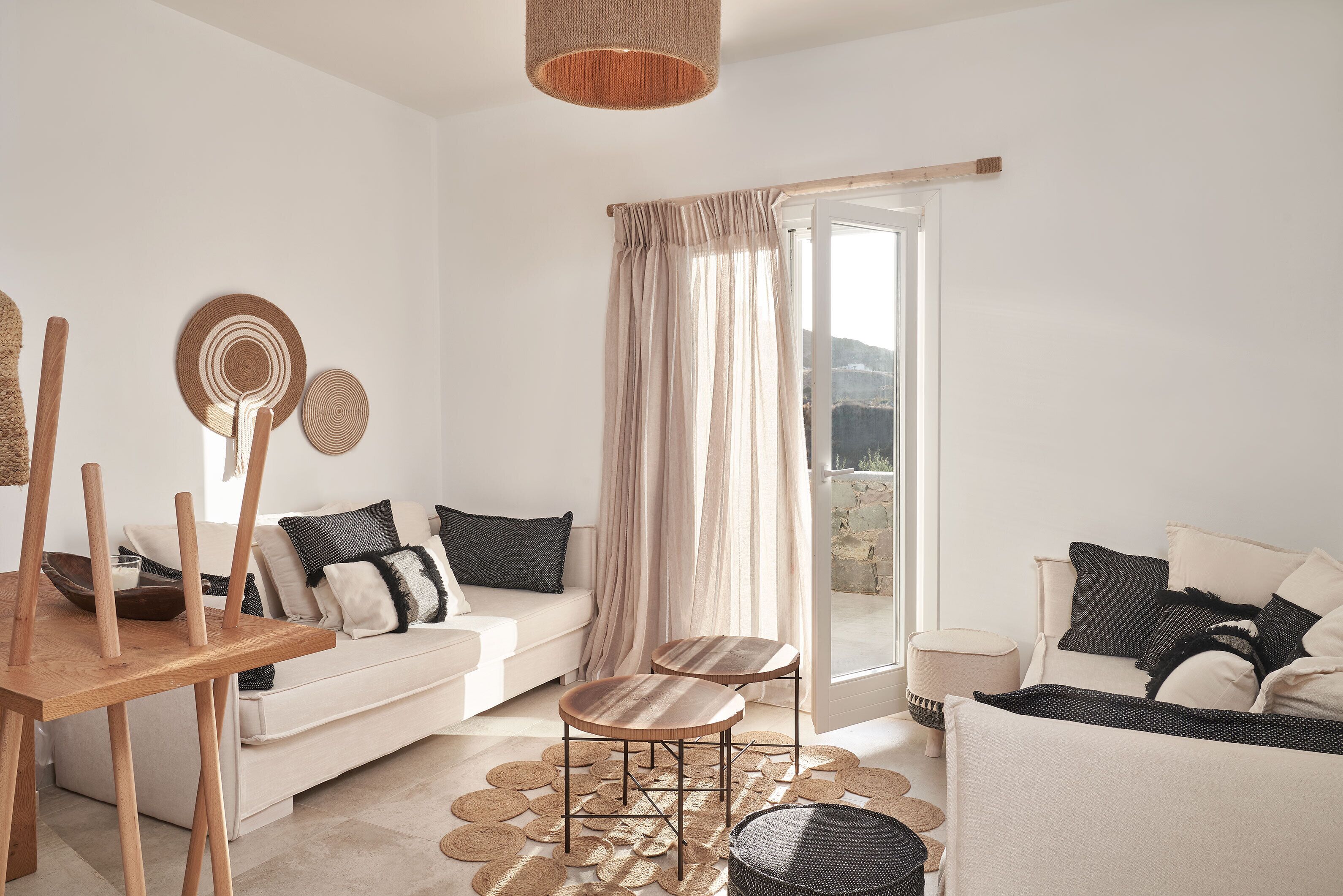 A modern living room with a white sofa, a wooden dining table, and a round coffee table. There is a large window with a view of the outdoors.