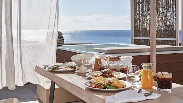 A table with breakfast items, a glass of orange juice, and a view of the ocean. - Volcano Luxury Suites Milos - Adults Only (Milos)