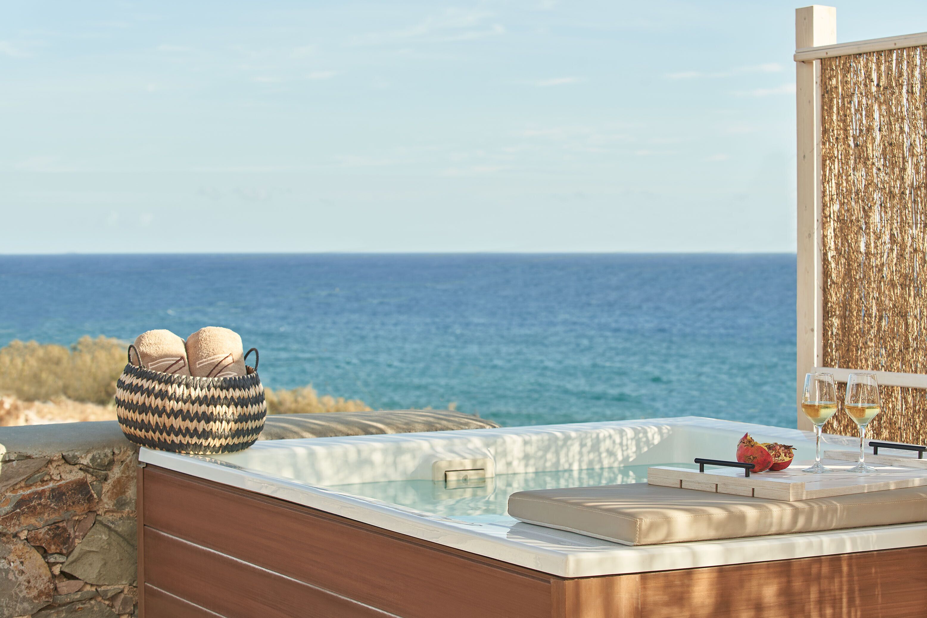 A hot tub with a view of the sea, a woven basket with cushions, and two glasses of wine on a wooden deck.