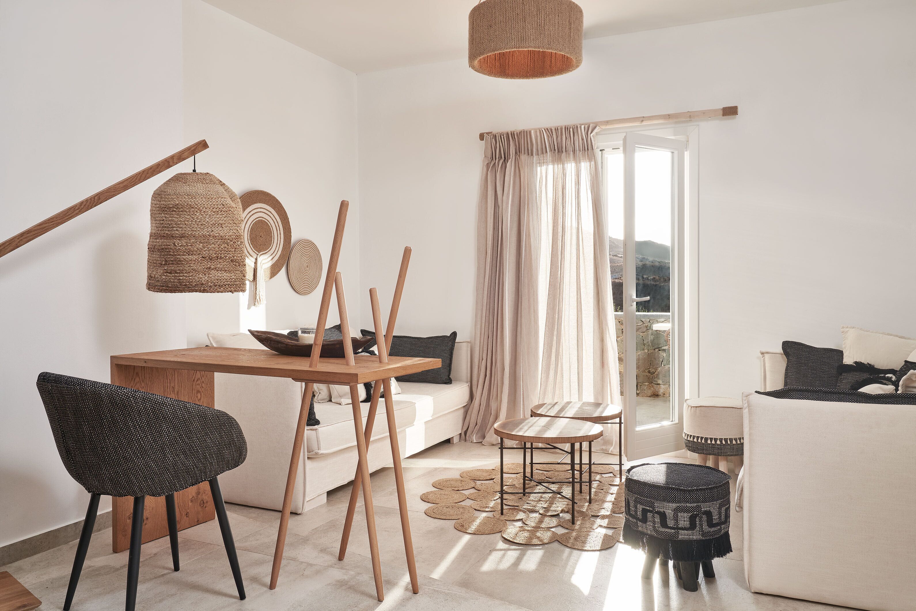 A modern living room with a wooden dining table, a woven chair, a sofa, and a view of the outdoors through a large window.