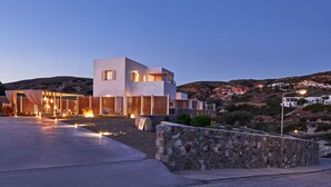 A modern building with a stone wall in the foreground, a paved area, and a mountainous backdrop. - Volcano Luxury Suites Milos - Adults Only (Milos)