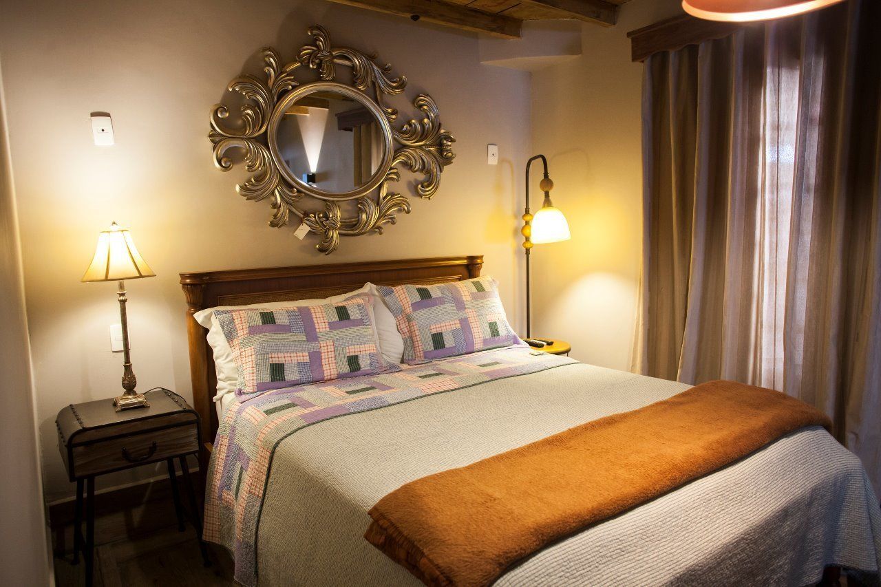 Un llit ben fet amb un llençol de tapet, una làmpada de nit, un mirall i una finestra amb cortines.