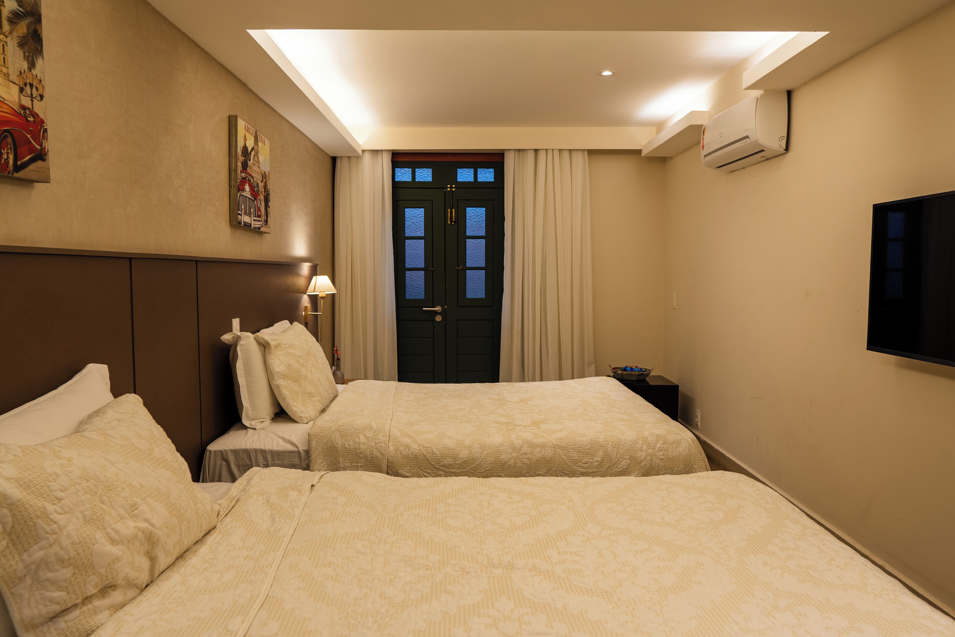 A hotel room with two beds, a door with curtains, an air conditioning unit, and a flat-screen TV on the wall.
