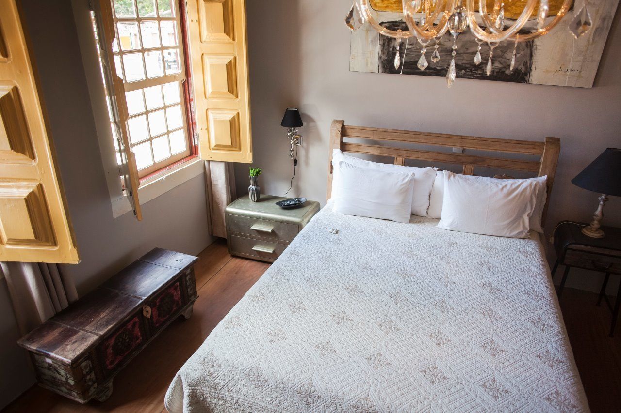 A bedroom with a wooden bed, white bedding, a nightstand, a lamp, a bench, and a window with open shutters.