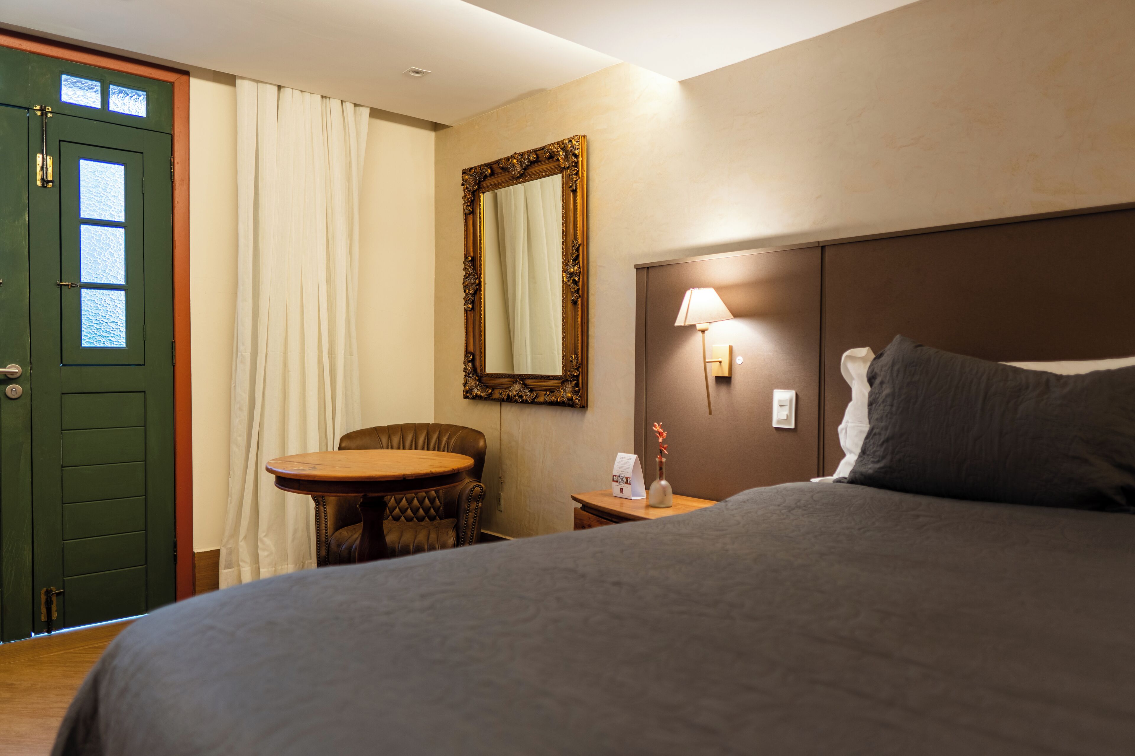 A hotel room with a bed, a round table, a chair, a mirror, and a lamp.
