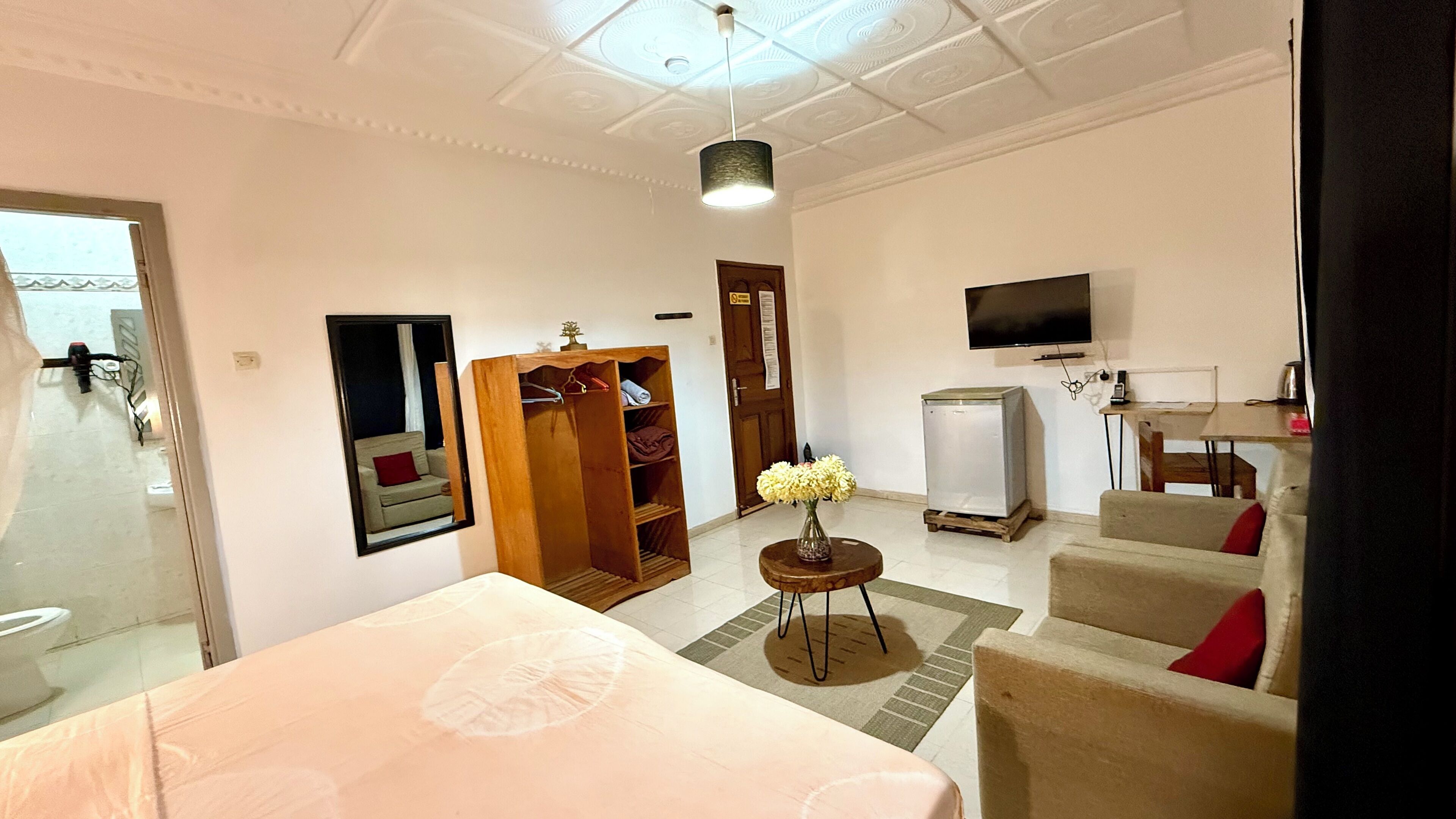 A hotel room with a bed, a wooden wardrobe, a small table with a vase, a refrigerator, a TV, and a sofa.