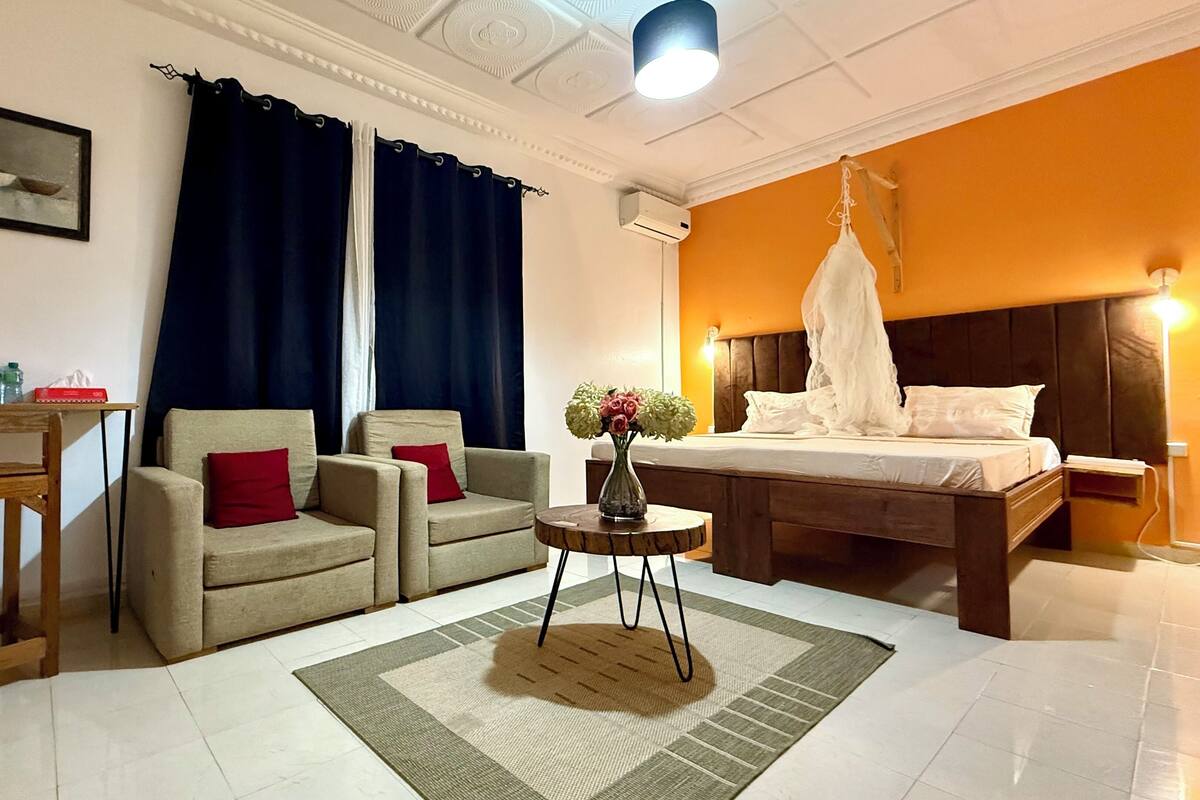 A hotel room with a bed, two armchairs, a small table with a vase, a painting on the wall, and a ceiling light.