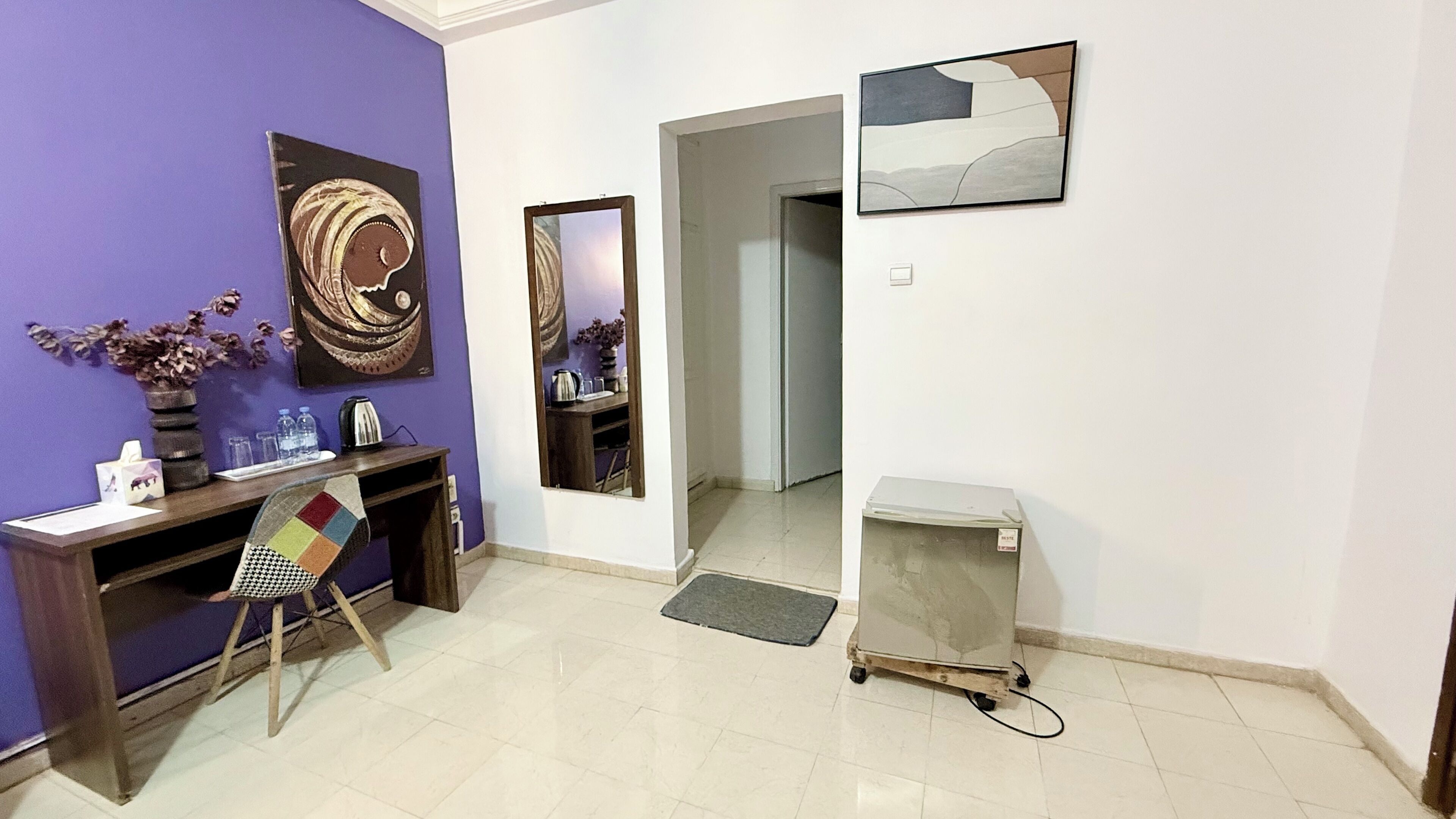 A room with a purple wall, a wooden desk, a chair, a small cabinet, and a mirror.