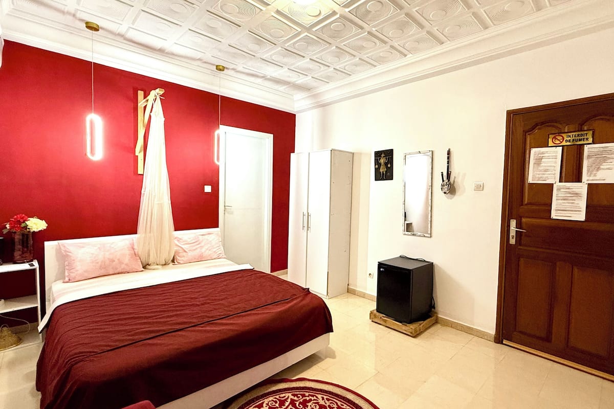 A bedroom with a red wall, a bed with a red bedspread, a white wardrobe, a wooden door with papers, and a ceiling with a decorative pattern.